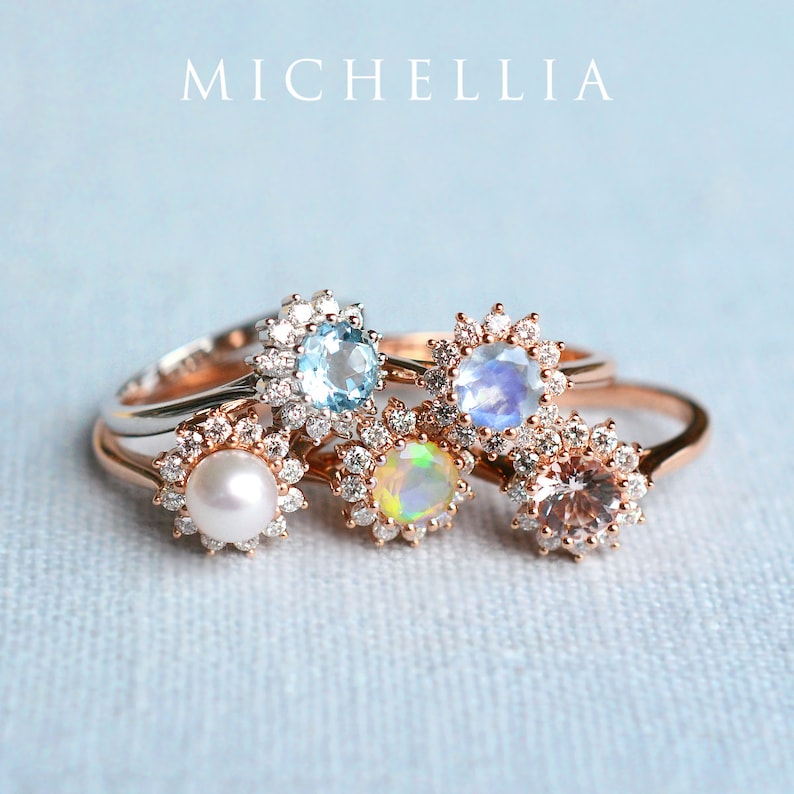 May include: Five rings with different gemstones set in a halo of diamonds. The rings are stacked on top of each other. The gemstones are aquamarine, moonstone, opal, morganite, and pearl.