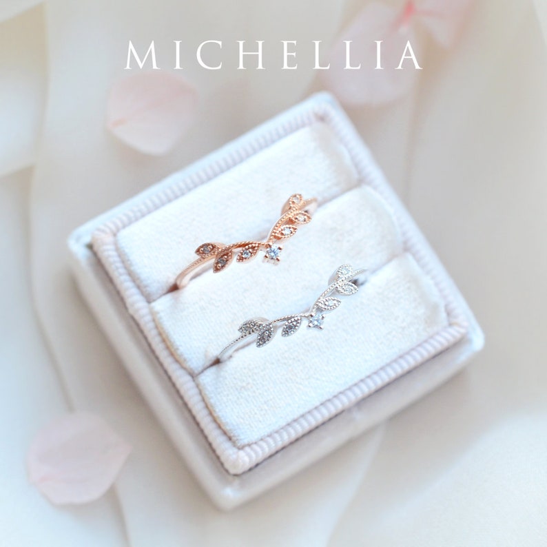 May include: Two delicate rings with a leaf design and small clear stones. The rings are in a white velvet ring box. The text "MICHELLIA" is visible above the box.