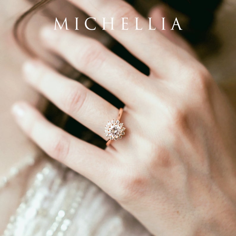 May include: A close-up of a rose gold ring with a round, clear diamond center stone surrounded by smaller diamonds. The ring is on a person's finger.