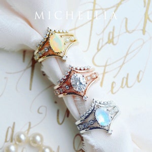 May include: Three gold rings with different gemstones. The top ring has an opal, the middle ring has a diamond, and the bottom ring has a moonstone. The rings are on a white fabric background.