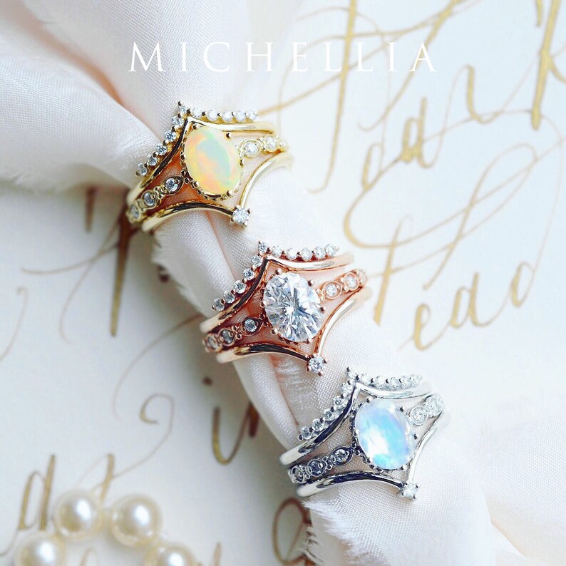 May include: Three gold rings with different gemstones. The top ring has an opal, the middle ring has a diamond, and the bottom ring has a moonstone. The rings are on a white fabric background.