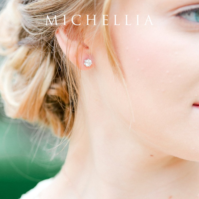 May include: A close-up of a person's ear with a small, round, clear gemstone earring. The earring is set in a silver or white gold setting. The background is blurred and out of focus.