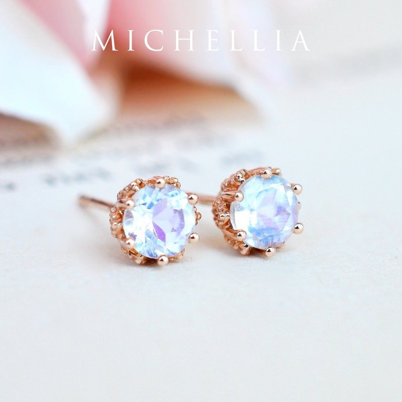 May include: A pair of rose gold earrings with a round, iridescent gemstone set in a delicate, ornate frame. The earrings are on a white background.