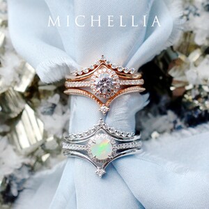 May include: Two delicate rings with a pointed crown design. The top ring features a round, clear gemstone surrounded by smaller stones. The bottom ring features a round, iridescent opal surrounded by smaller stones. Both rings are set in a silver band.