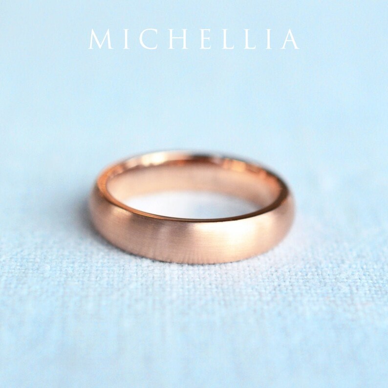 May include: A rose gold wedding band with a brushed finish. The ring is set against a light blue background. The word "MICHELLIA" is printed in white at the top of the image.