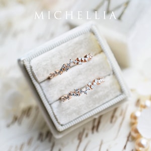 Pu&ograve; includere: Due delicati anelli in oro rosa con un designo di diamanti sparsi, esposti in una scatola per anelli in velluto bianco. Gli anelli sono disposti a scalare, mostrando i dettagli intricati delle montature di diamanti.