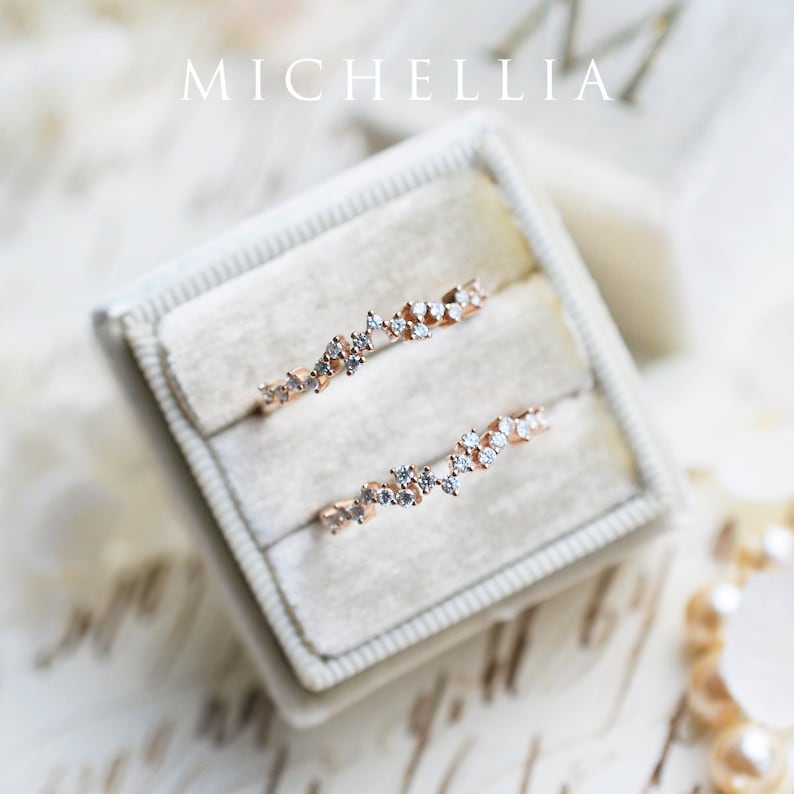 Pu&ograve; includere: Due delicati anelli in oro rosa con un designo di diamanti sparsi, esposti in una scatola per anelli in velluto bianco. Gli anelli sono disposti a scalare, mostrando i dettagli intricati delle montature di diamanti.