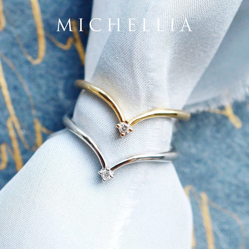May include: Two gold and silver V-shaped rings with a small diamond at the point of the V. The rings are on a light blue fabric background.