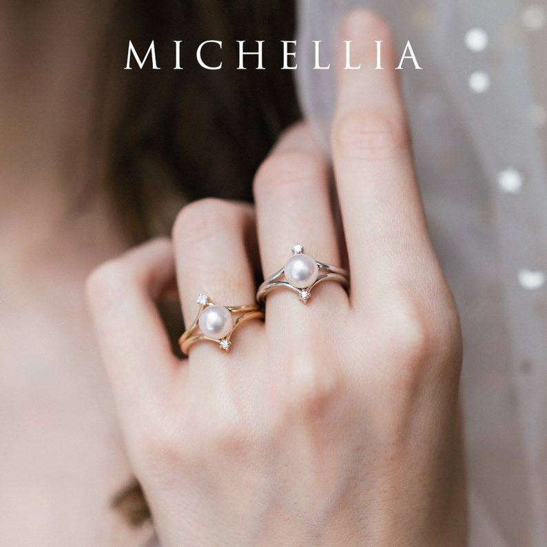 May include: Two gold and silver rings with a pearl and small diamonds. The rings are on a person's finger.