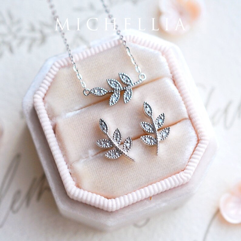 Op de afbeelding: Een set zilveren sieraden met een delicaat bladontwerp met glinsterende heldere stenen. De ketting en oorbellen worden gepresenteerd in een roze fluwelen doosje.