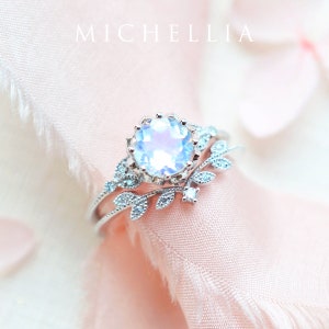 May include: A silver ring set with a large, iridescent moonstone surrounded by smaller stones. The ring is accented with a delicate leaf design.