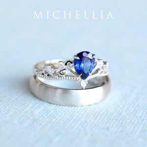May include: A pair of wedding rings. The top ring is silver with a pear-shaped, deep blue gemstone and small accent stones. The second ring is a simple, brushed silver band. The word "MICHELLIA" is at the top.