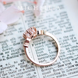 May include: A rose gold engagement ring with a large, clear centre stone surrounded by smaller stones. The ring is set on a page of a book with the word "Flower" visible.