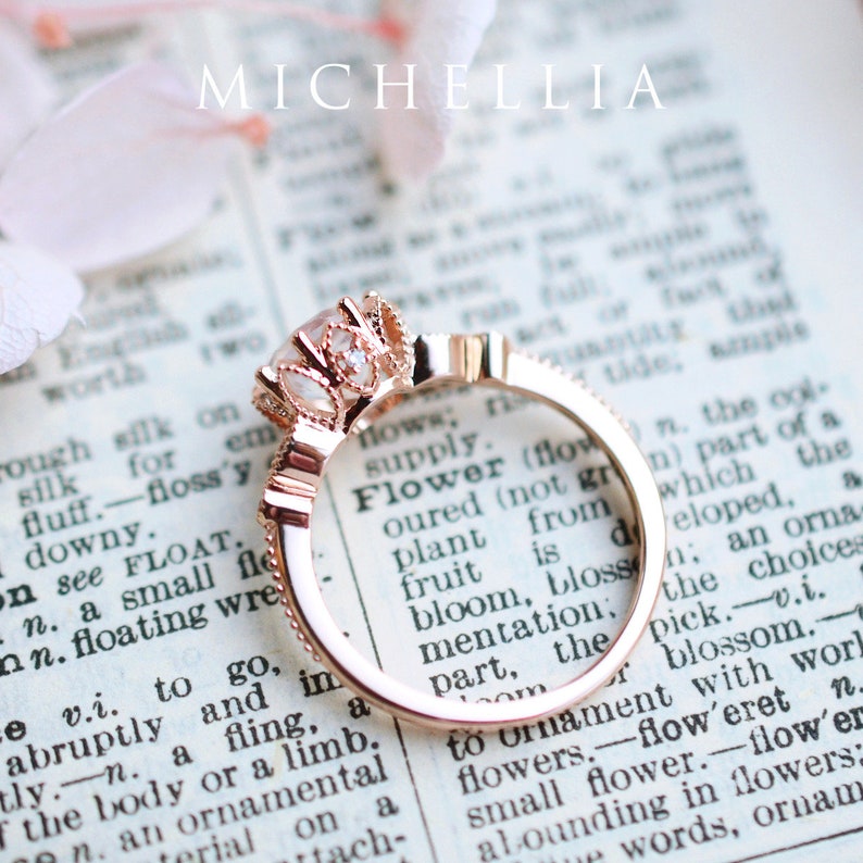 May include: A rose gold engagement ring with a large, clear centre stone surrounded by smaller stones. The ring is set on a page of a book with the word "Flower" visible.