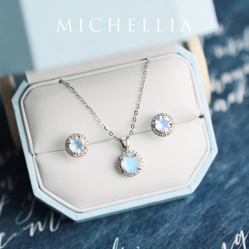 Pu&ograve; includere: Un set di collana e orecchini in argento con un ciondolo in pietra di luna e orecchini abbinati. Le pietre sono circondate da un alone di diamanti pi&ugrave; piccoli. I gioielli sono esposti in una scatola blu chiaro con la scritta "MICHELLIA" stampata sulla parte superiore.