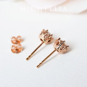 May include: A pair of rose gold stud earrings with a clear stone set in a detailed, textured setting. The earrings have a post back.