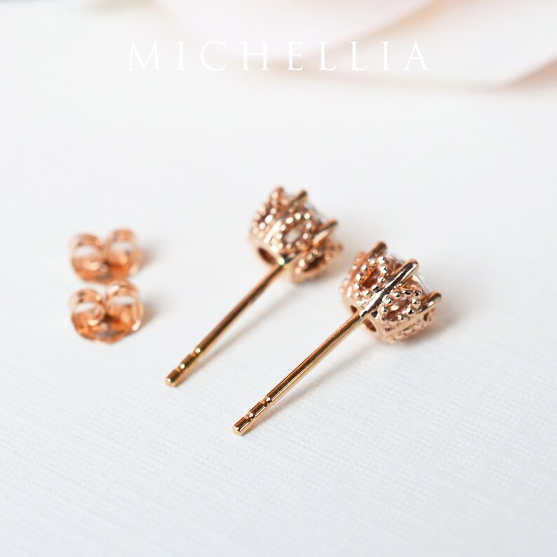 May include: A pair of rose gold stud earrings with a clear stone set in a detailed, textured setting. The earrings have a post back.