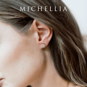 May include: A pair of rose gold stud earrings with a round, clear stone surrounded by smaller stones. The earrings are worn in a person's ear.