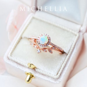 May include: A delicate rose gold ring set with a white opal gemstone surrounded by a halo of small clear stones. The ring is paired with a matching band featuring a delicate leaf design.