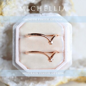 May include: Two gold-toned rings with a small diamond accent at the point of the V-shaped design. The top ring has a smooth finish, while the bottom ring has a milgrain finish. The rings are displayed in a white velvet ring box. "MICHELLIA" and "SMOOTH FINISH OPTION" are written above the rings, and "MILGRAIN FINISH OPTION" is written below the rings.