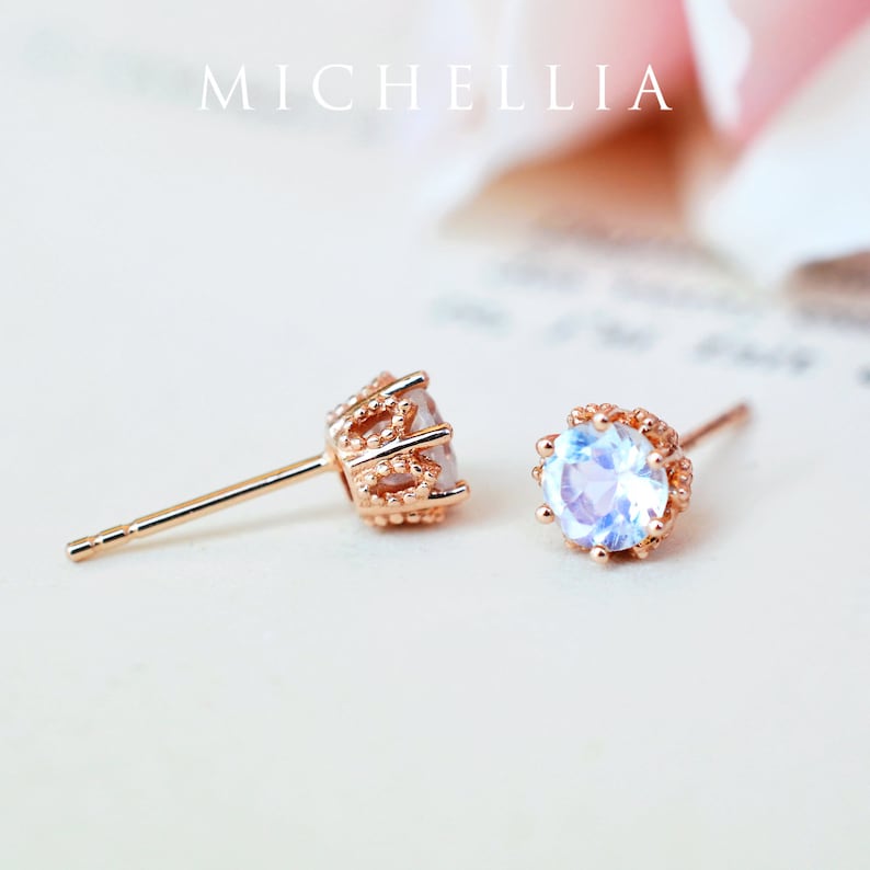 May include: A pair of rose gold earrings with a round, iridescent gemstone set in a delicate crown-shaped setting. The earrings have a post back.