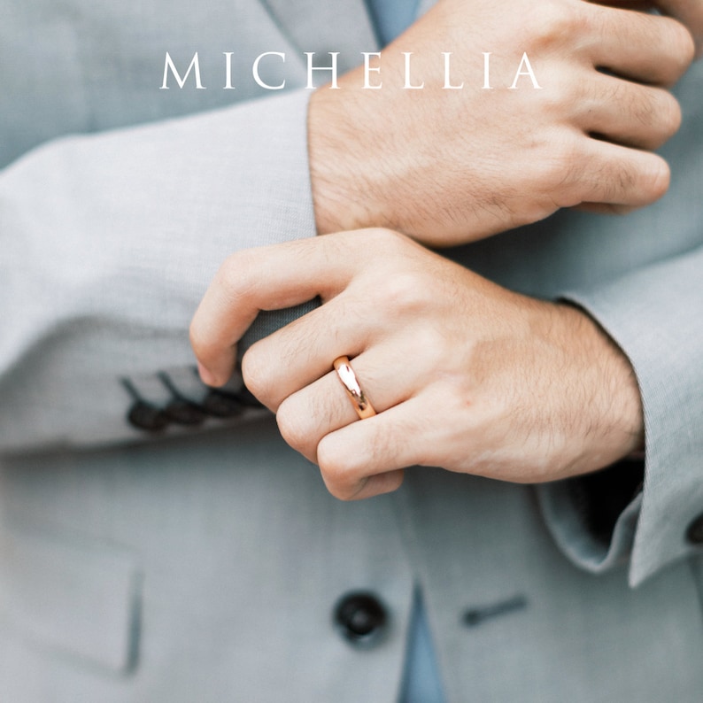 May include: A rose gold ring is displayed on a person's finger, set against a light gray suit. The ring has a smooth, polished finish. The word "MICHELLIA" is printed in white at the top of the image.
