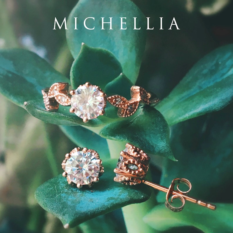 May include: A rose gold engagement ring and matching earrings with a large, clear, round gemstone. The ring features a delicate leaf design and the earrings have a similar design. The jewelry is displayed on a green leafy background.