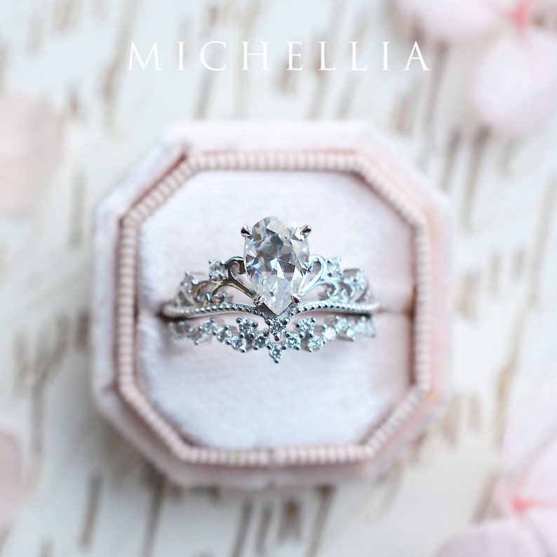 Pu&ograve; includere: Un anello di fidanzamento con diamante a forma di pera con una delicata montatura a corona. L'anello &egrave; in una scatola per anelli in velluto rosa. Il testo "MICHELLIA" &egrave; visibile sopra la scatola dell'anello.