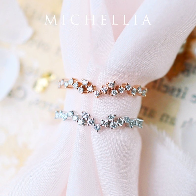 Pu&ograve; includere: Due anelli delicati con un design a forma di onda incastonati con piccoli diamanti scintillanti. Gli anelli sono su uno sfondo di tessuto rosa morbido.