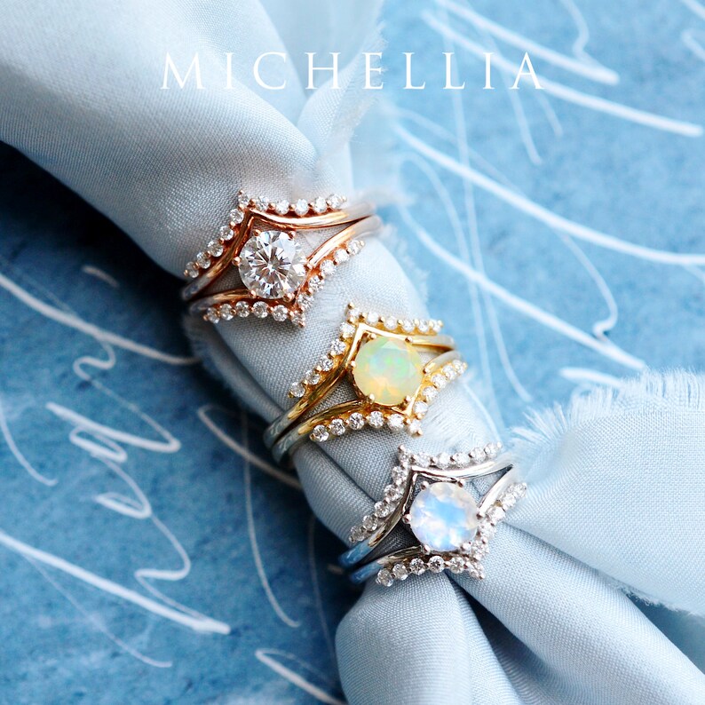 May include: Three gold rings with different gemstones set in a V-shaped design. The rings are on a light blue fabric background. The text "MICHELLIA" is visible in the top center of the image.