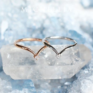 May include: Two delicate V-shaped rings with a single diamond accent at the point of the V. The rings are shown on a clear crystal surface.