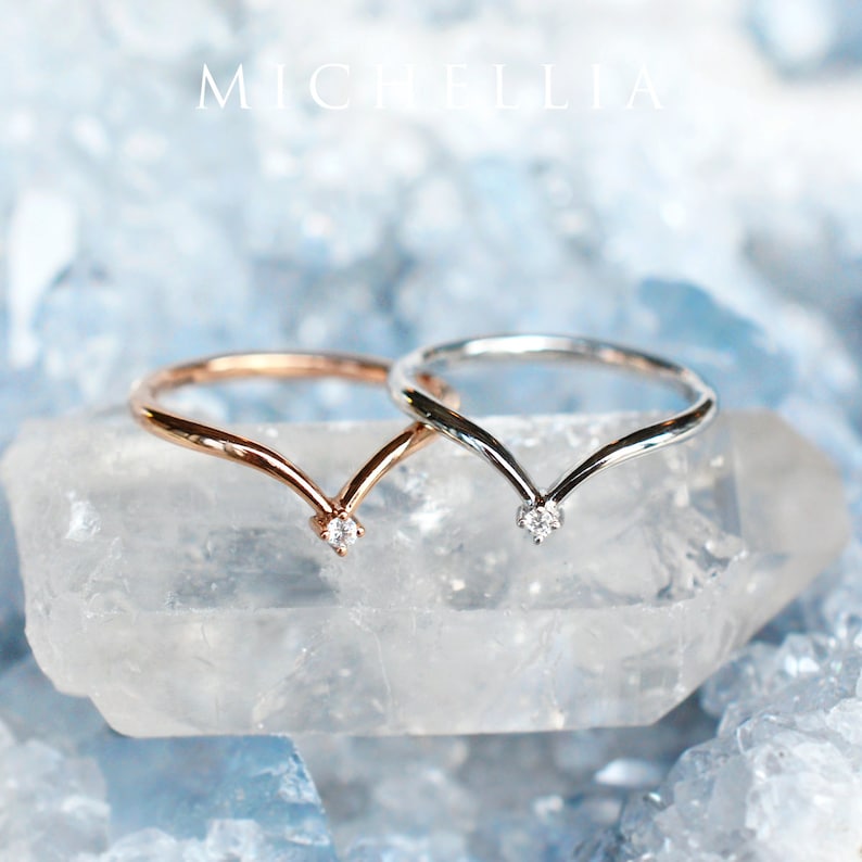 May include: Two delicate V-shaped rings with a single diamond accent at the point of the V. The rings are shown on a clear crystal surface.