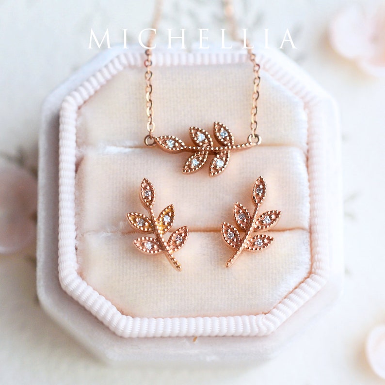 Op de afbeelding: Een set rose gouden sieraden met een delicaat bladontwerp met glinsterende heldere stenen. De ketting en oorbellen worden gepresenteerd in een roze fluwelen doos.
