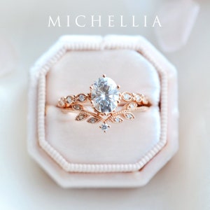 May include: A rose gold engagement ring set with a large oval-shaped center stone and a delicate band with small stones and leaf accents. The ring is in a pink velvet ring box.