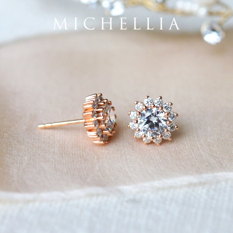May include: A pair of rose gold stud earrings with a large, clear, round gemstone surrounded by smaller, clear gemstones. The earrings are on a white background.