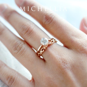 K&ouml;nnte beinhalten: Nahaufnahme einer Frauenhand mit einem Verlobungsring aus Ros&eacute;gold mit einem gro&szlig;en, runden, klaren Diamanten und einem zarten, verzierten Ehering aus Ros&eacute;gold.