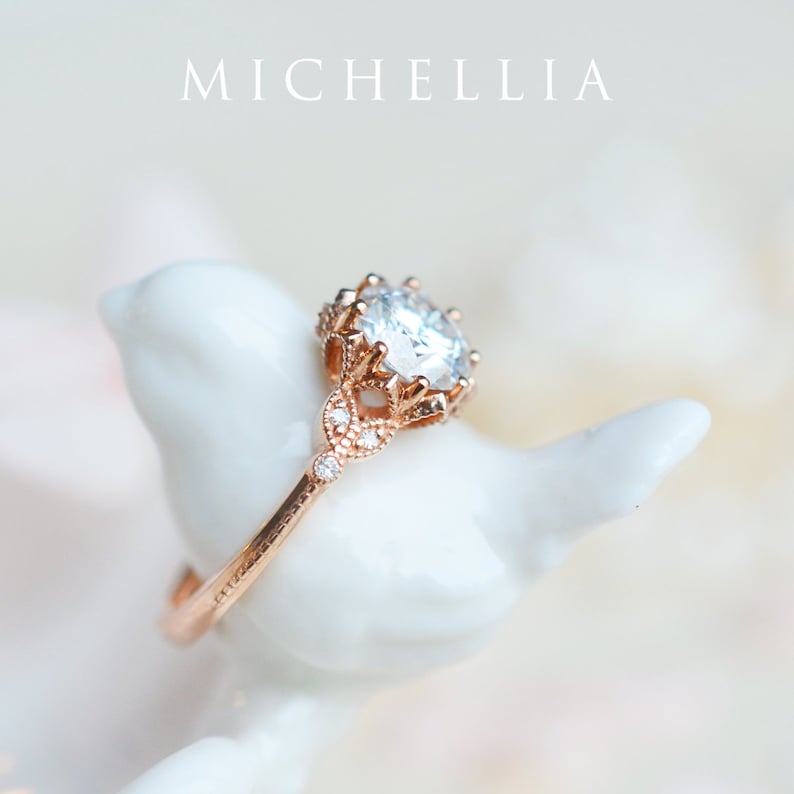 May include: A rose gold engagement ring featuring a large, round, clear centre stone and smaller stones set in a delicate, ornate design.