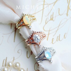 May include: Three gold rings with different gemstones. The top ring has an opal, the middle ring has a diamond, and the bottom ring has a moonstone. The rings are on a white fabric background.