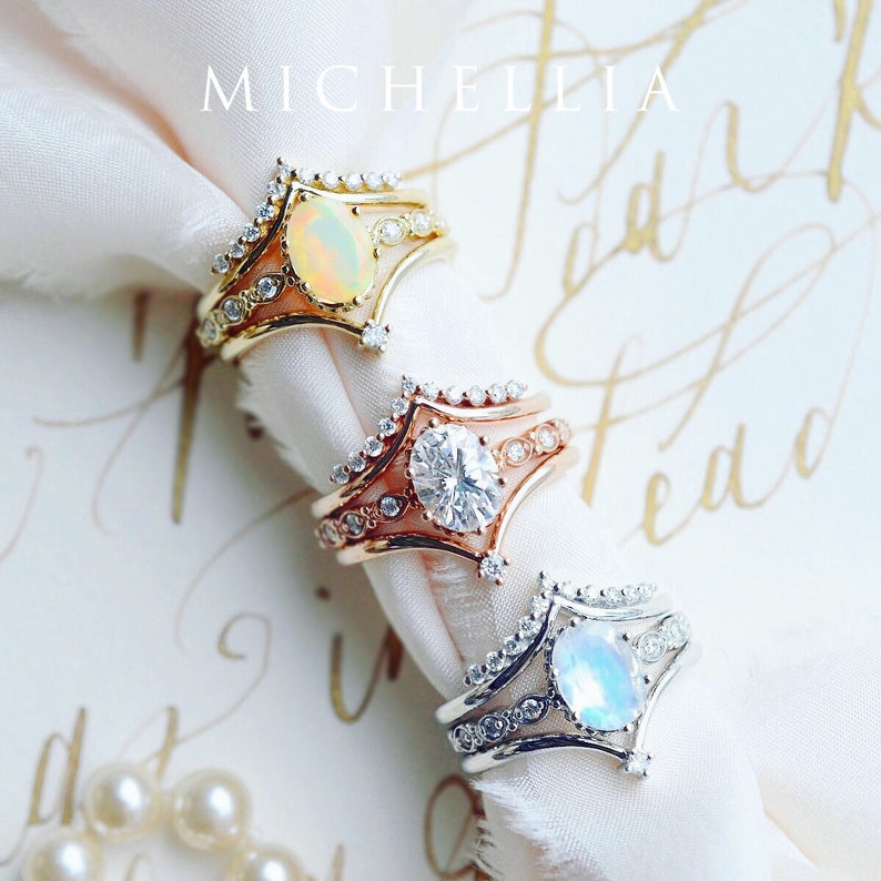 May include: Three gold rings with different gemstones. The top ring has an opal, the middle ring has a diamond, and the bottom ring has a moonstone. The rings are on a white fabric background.