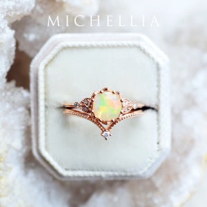 May include: A delicate rose gold ring set with a large opal gemstone and smaller diamonds. The ring is in a white velvet ring box. The box has the word "MICHELLIA" printed on the top.