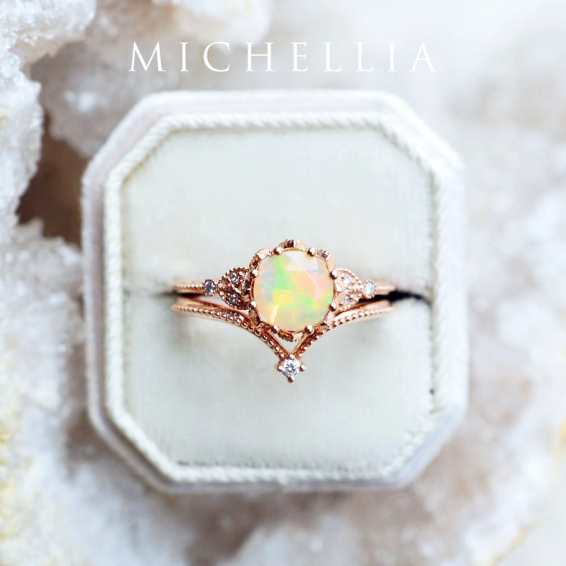May include: A delicate rose gold ring set with a large opal gemstone and smaller diamonds. The ring is in a white velvet ring box. The box has the word "MICHELLIA" printed on the top.