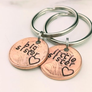 May include: Two copper keychains with silver key rings. One keychain says "big sister" with a heart and the other says "little sister" with a heart.
