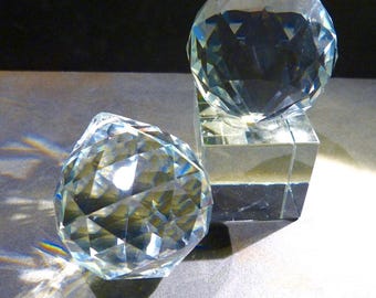 Puede incluir: Dos esferas de cristal transparente con superficies facetadas. Una esfera descansa sobre una base cuadrada transparente, la otra sobre una superficie gris. Las esferas reflejan la luz, creando un efecto brillante. Esferas decorativas de cristal.