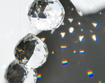 Puede incluir: Tres esferas de cristal transparente colgando sobre un fondo blanco. Las esferas están facetadas, reflejando la luz y creando un efecto de prisma arcoíris sobre una superficie gris oscuro. Las esferas están suspendidas por hilos finos, casi invisibles.