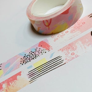 May include: A roll of decorative washi tape with a pastel abstract design. The tape features pink, yellow, and blue brushstrokes, black dots, and lines. Two strips of the tape are unrolled, showing the pattern.