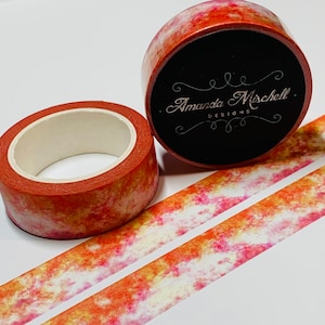 May include: Two rolls of washi tape with a watercolor design in shades of orange, yellow, and pink. The tape is unrolled on a white surface. The roll of tape in the foreground has the text "Amanda Mischell Designs" on the label.
