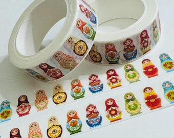 Puede incluir: Dos rollos de cinta adhesiva blanca con un patrón repetitivo de coloridas muñecas rusas. Cada muñeca está ilustrada con detalles florales y geométricos distintos en tonos rojo, amarillo, verde y azul, dispuestos en una línea horizontal.
