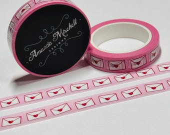 May include: Pink washi tape with a repeating pattern of white envelopes with red hearts. The tape is rolled up on a pink spool with the text "Amanda Mischell Designs" printed on the spool.