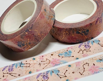 May include: Two rolls of decorative washi tape with a celestial theme. The tape features constellations in gold, with a watercolor background of pink, purple, and blue. Two strips of tape are unrolled, showing a floral and constellation pattern.