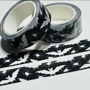 May include: Two rolls of decorative washi tape with a Halloween theme. The tape features a black background with a pattern of white bats and spiders. A strip of the tape is unrolled, showing the repeating design.